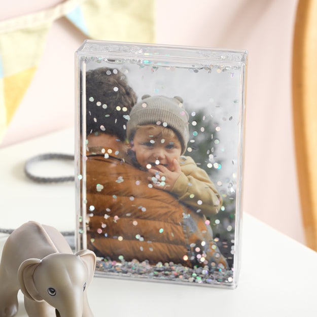 Clear photo frame with glitter filled with a photo of a child, placed on a surface.