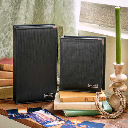 Two black slip-in photo albums with 'PHOTOS' text on the front, displayed on a wooden surface alongside a candlestick and books.