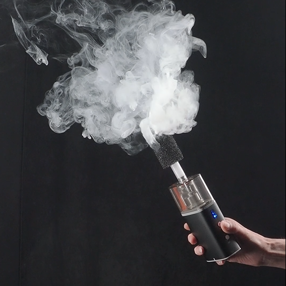 Hand holding a fog machine device emitting white smoke against a black background